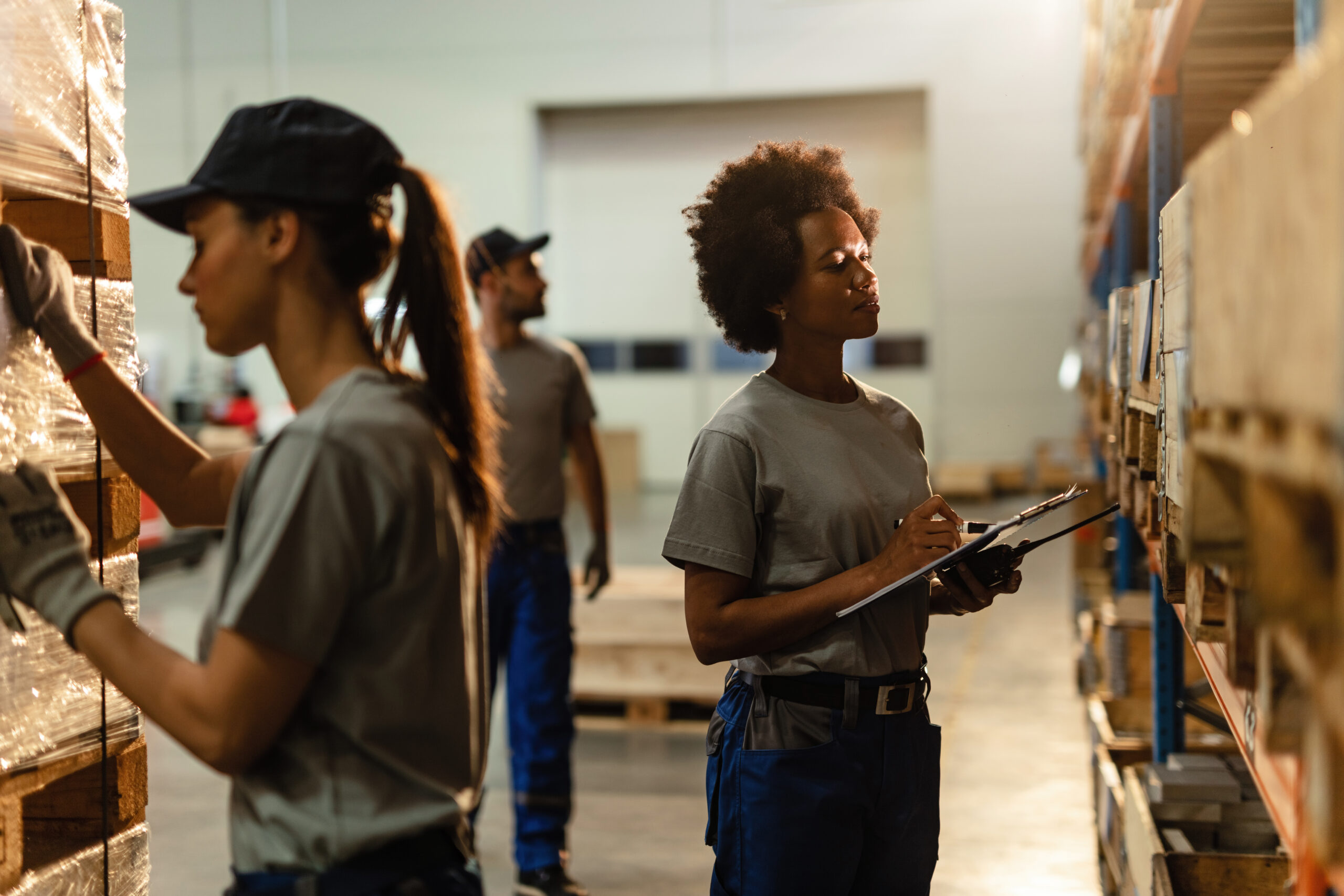 Black female warehouse worker going through shipment list while checking stock in industrial storage compartment.